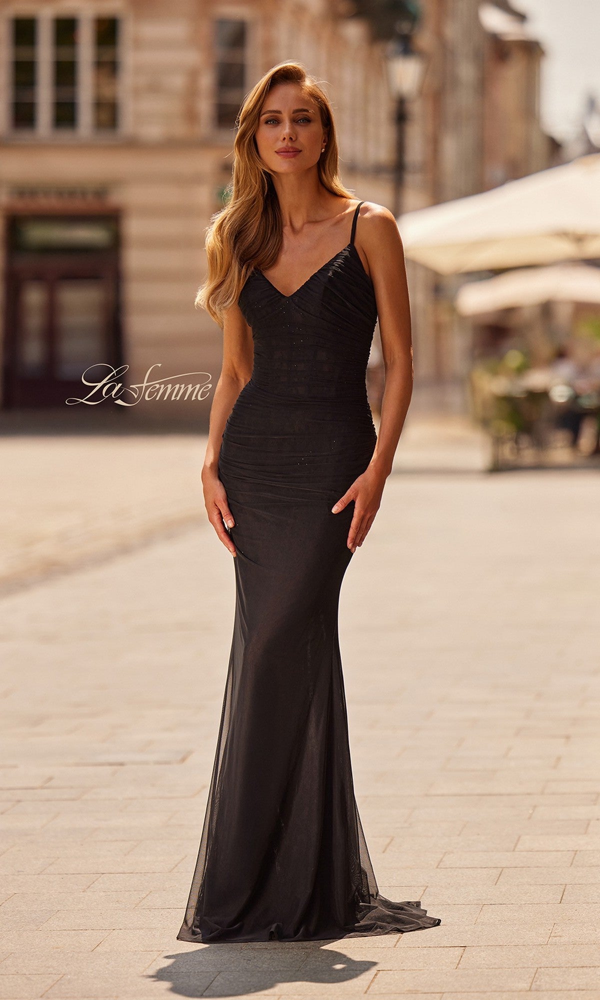 A woman stands on a sunlit cobblestone street in the La Femme 33411 Long Prom Dress, a fitted sleeveless black gown featuring a v-neckline and thin straps, with blurred buildings and outdoor seating in the background.